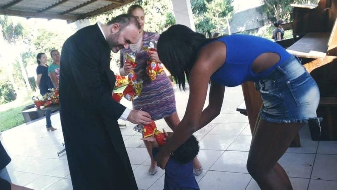 Voluntária e sacerdote entregam presente a uma criança